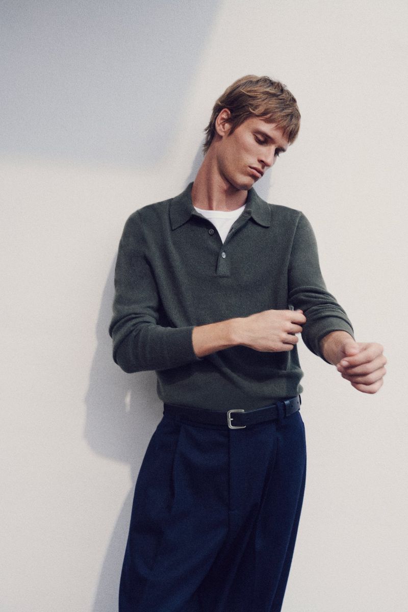 Adult man in olive green polo shirt and navy trousers adjusting his sleeve in a neutral indoor setting with soft, diffused lighting.