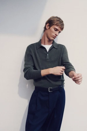Adult man in olive green polo shirt and navy trousers adjusting his sleeve in a neutral indoor setting with soft, diffused lighting.