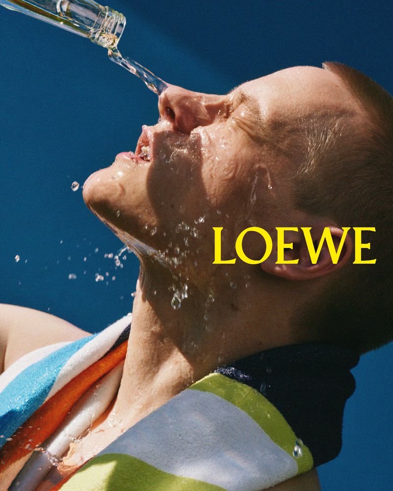 Close-up of a man's face as water is poured from a clear glass bottle in an outdoor setting, with 'LOEWE' text overlay.