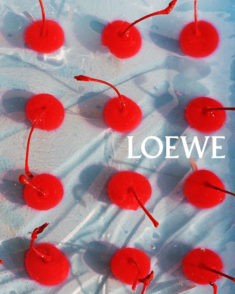 Overhead shot of bright red cherries arranged symmetrically on a light blue surface, with the word LOEWE visible.