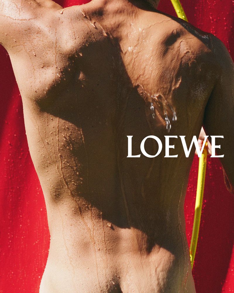 Torso of adult male with water droplets and yellow tube against a red background with the Loewe logo visible, studio lighting.