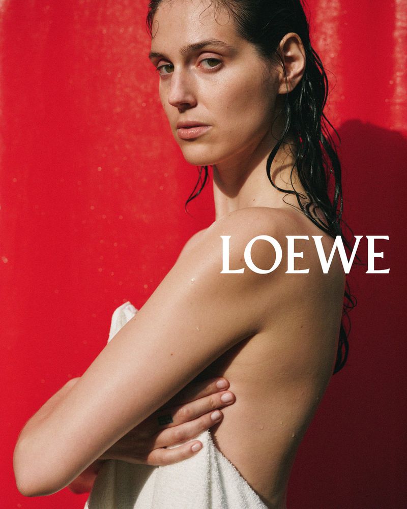 Striking portrait of a woman with wet hair, partially covered by a towel, against a vibrant red background, featuring the LOEWE logo.