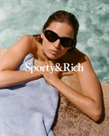 Woman wearing sunglasses and wrapped in a towel, resting against tan stone with turquoise water background. Sporty & Rich branding visible.