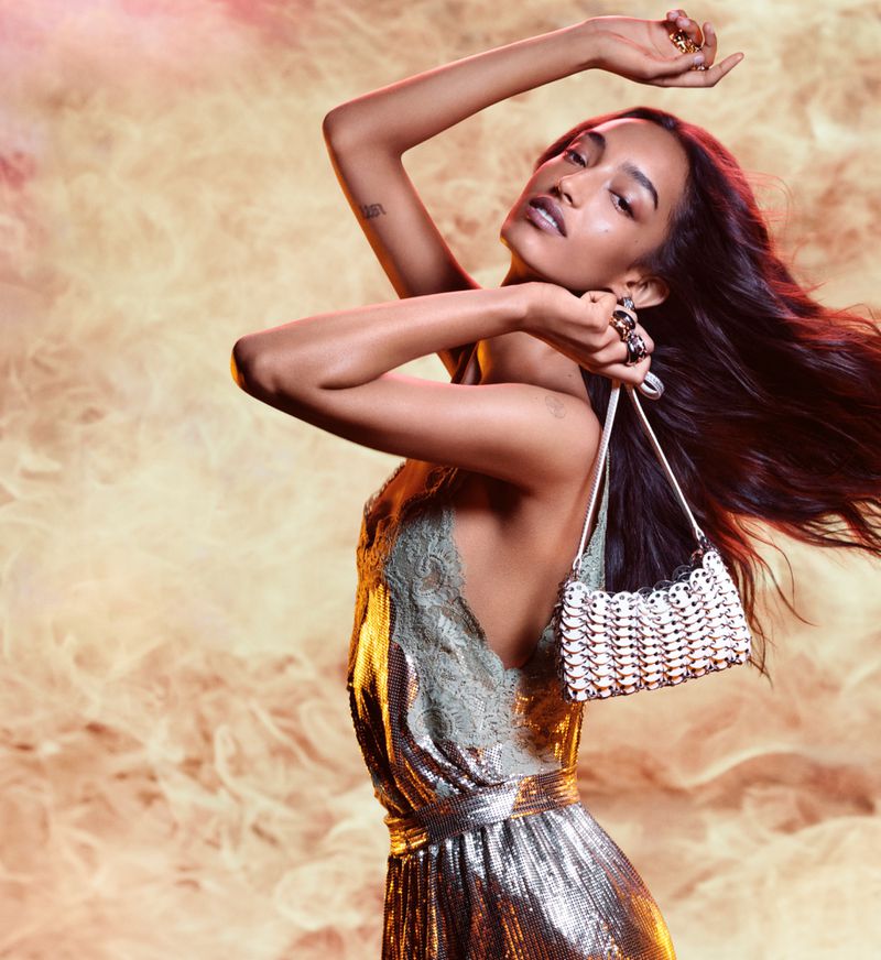 Fashionable adult woman poses in a glittering dress with a purse against a smoky background, showcasing style and elegance.