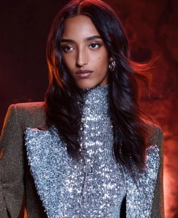A woman with dark hair wearing a sparkly top and jacket poses in a studio with a red background, staring intensely at the camera.