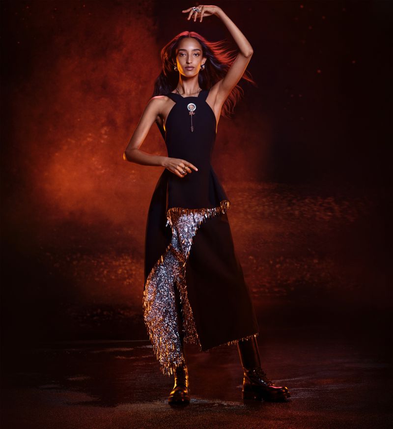 Fashionable woman in black dress with silver accents strikes a pose under orange lighting in a studio fashion editorial shot.