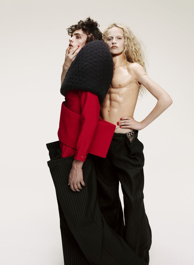 Striking fashion image featuring two androgynous adults, one in red and black, the other shirtless in black pants, in a bright studio.