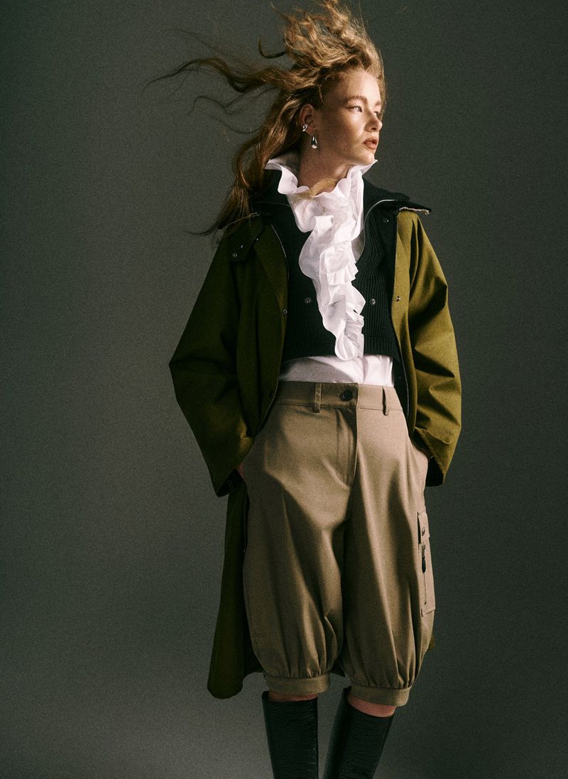 Stylish woman in olive coat, white ruffled top, balloon trousers, and boots, posing against a plain grey backdrop in a studio shot.