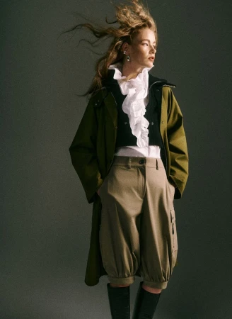 Stylish woman in olive coat, white ruffled top, balloon trousers, and boots, posing against a plain grey backdrop in a studio shot.