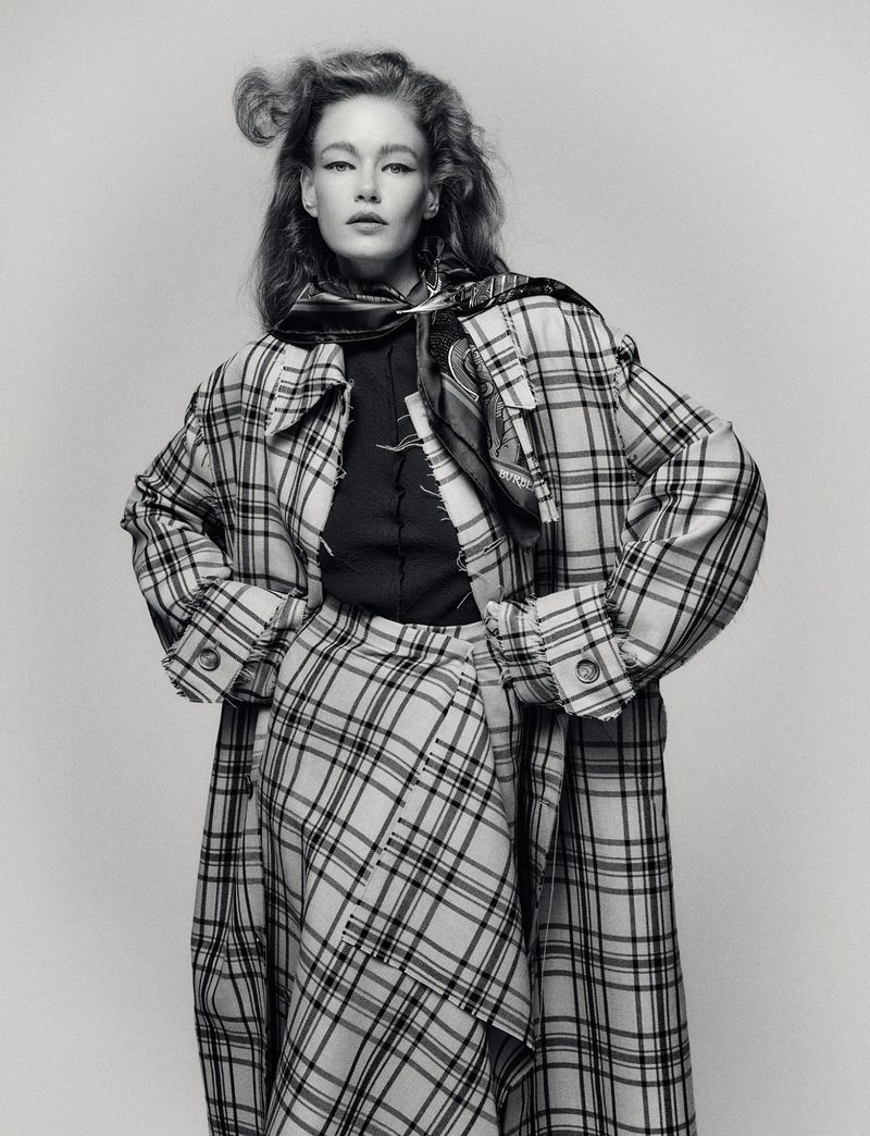 Monochrome portrait of a stylish adult woman wearing a plaid coat and skirt, posing indoors against a neutral backdrop.
