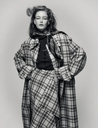 Monochrome portrait of a stylish adult woman wearing a plaid coat and skirt, posing indoors against a neutral backdrop.