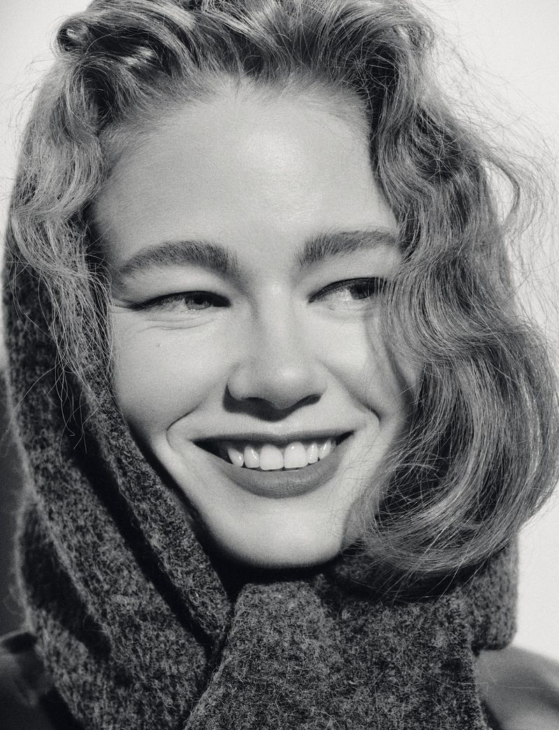Close-up portrait of a smiling woman with curly hair wearing a dark-colored, textured headscarf in a black and white photo.