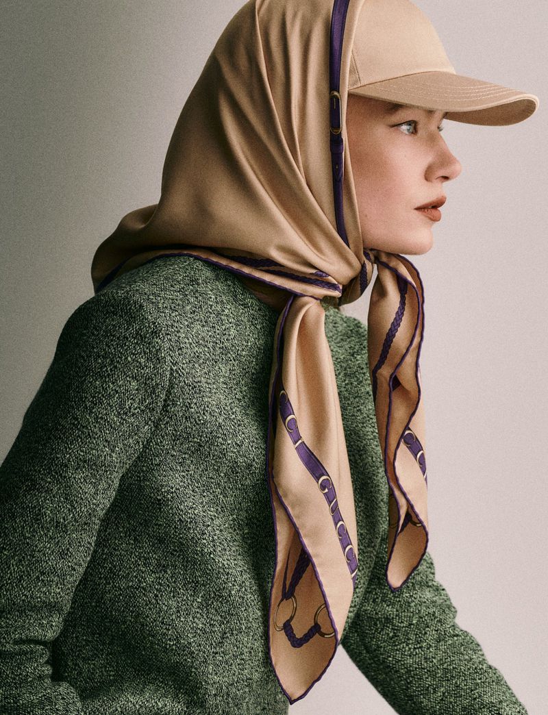 Portrait of a young woman with a tan baseball cap and patterned scarf, wearing a tweed sweater and lipstick in an indoor setting.