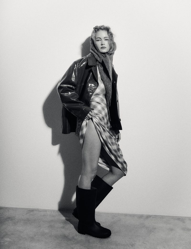 Black and white fashion shot of a woman in a shiny jacket, scarf, plaid dress, and boots posing in a minimalistic photo studio.