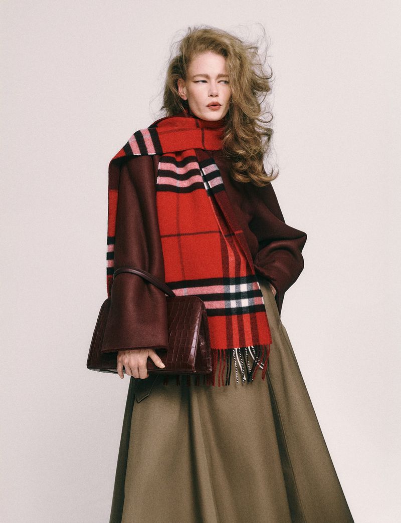 Fashion shot of woman in maroon coat, tan skirt, and red plaid scarf holding a dark brown purse, posed against a plain background.