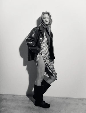 Model Hollie-May Saker wearing a glossy jacket, draped plaid dress, headscarf, and knee-high boots in a black and white studio portrait.