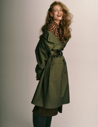 Model wearing an olive green trench coat and plaid shirt smiling against a neutral background for Vogue Greece