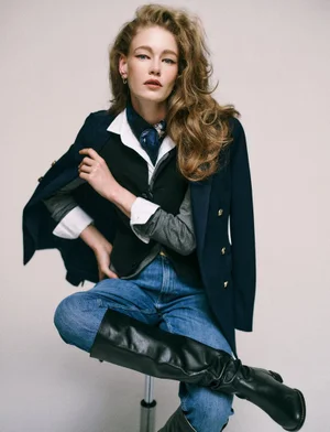 Model Hollie May Saker wearing a navy blazer, layered vest, white shirt, and black boots against a plain backdrop.