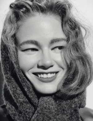 Close-up portrait of a smiling woman with wavy hair wearing a textured dark hooded scarf against a plain background.