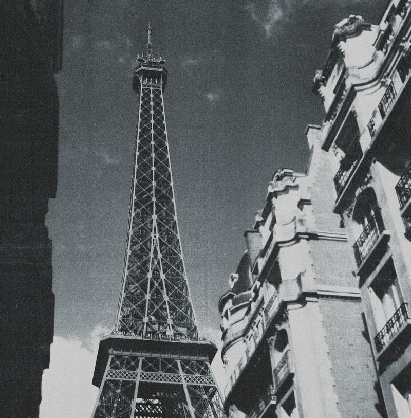 Low-angle shot of the Eiffel Tower in Paris, France, framed by classic urban architecture, in black and white with a cloudy sky.
