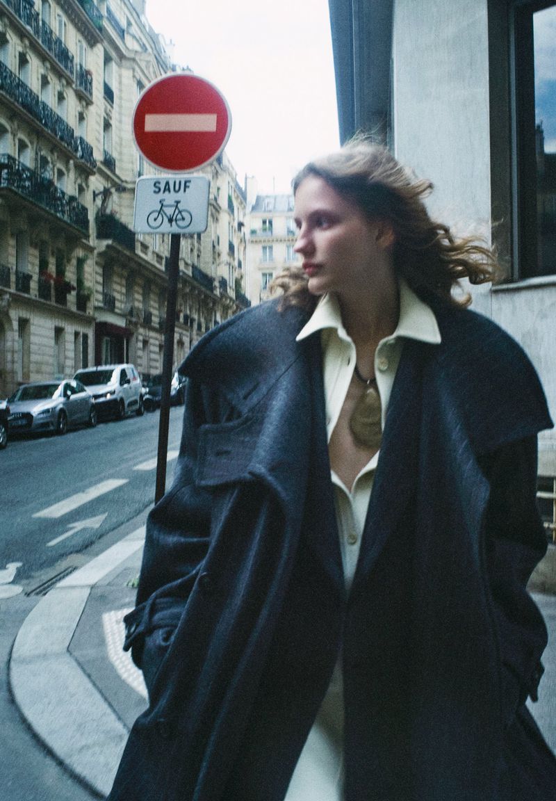 A woman in a coat standing on a city street with European architecture, a signpost, and cars in the background on a cloudy day.