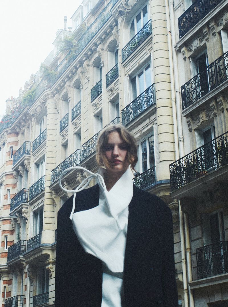 Woman in oversized blazer and draped white shirt posing in front of classic architecture, overcast daylight, medium shot.