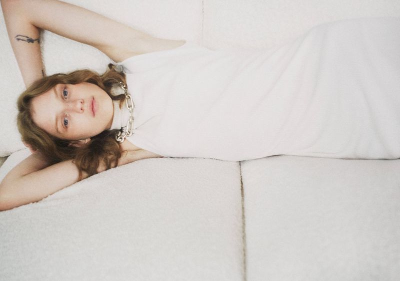 Red-haired woman in a white dress lies on a white couch, gazing forward with a silver chain necklace, relaxed pose, and soft lighting.