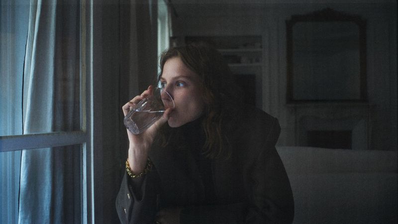 Woman with curly hair drinks water from a glass while standing by a window with white curtains in a dimly lit indoor setting.