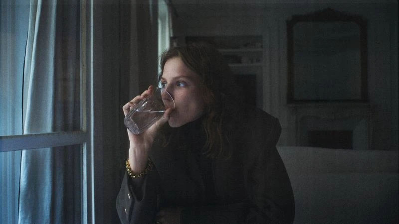 Woman with curly hair drinks water from a glass while standing by a window with white curtains in a dimly lit indoor setting.