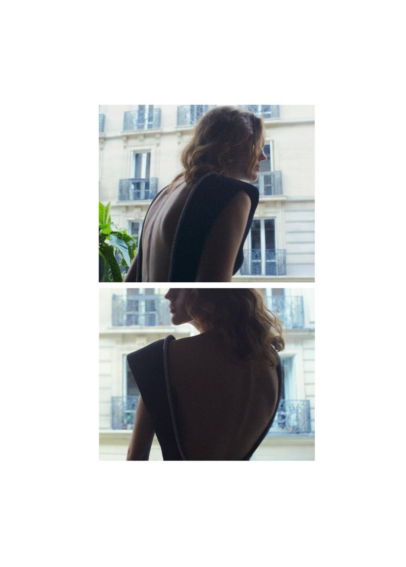 Adult female model poses in front of building wearing a dark, sleeveless dress, looking to the side in natural light in an outdoor environment.