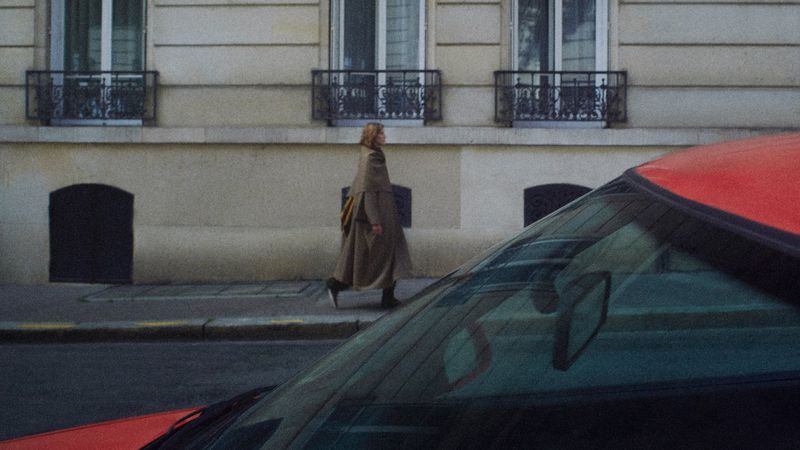 Woman in a coat walks along a city street past a red car. Urban scene with a pedestrian and architecture in the background.