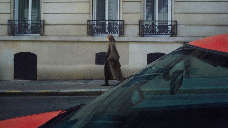 Model Nadine Kirilova walking down a street in a long brown coat, viewed from behind the windshield of a red car.