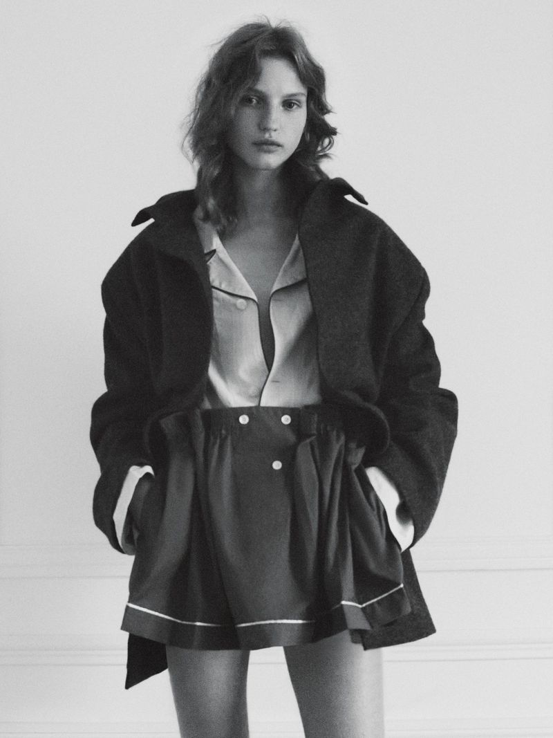 A fashion portrait showcases a young woman in a stylish, oversized coat and shorts, set against a neutral backdrop in black and white.