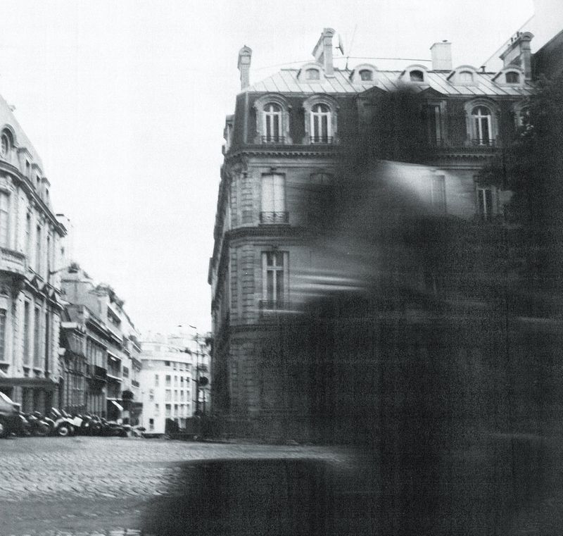 Monochrome urban scene with Parisian architecture, cobblestone street, and a blurred foreground element suggesting movement, creating a dynamic feel.