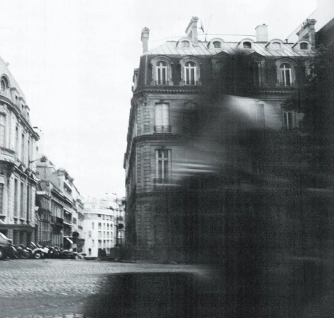 Monochrome urban scene with Parisian architecture, cobblestone street, and a blurred foreground element suggesting movement, creating a dynamic feel.