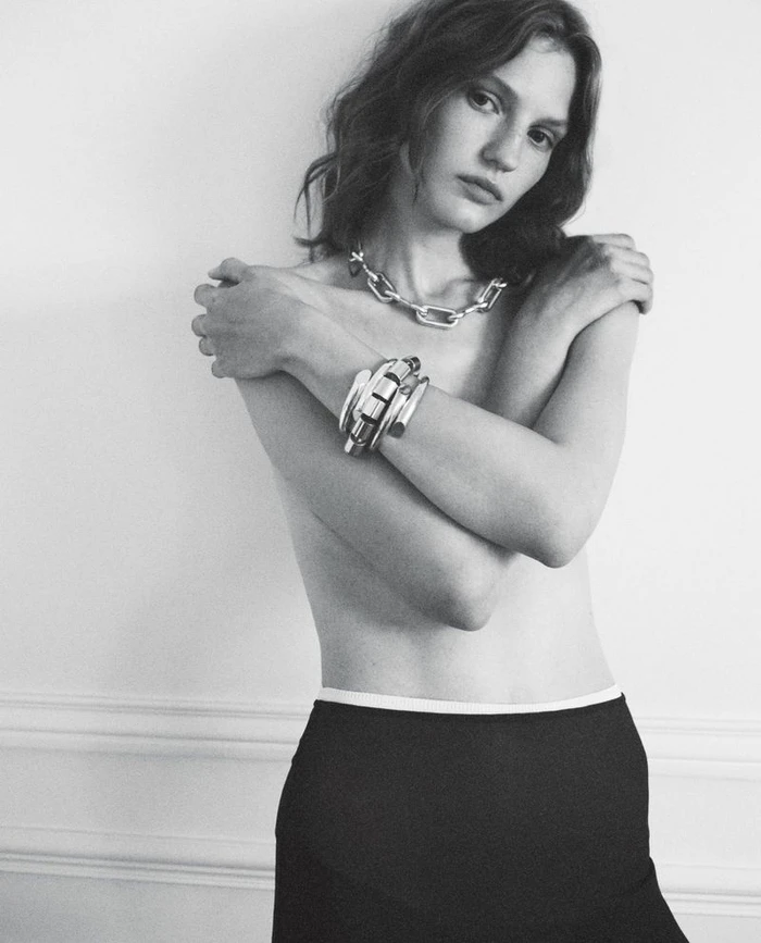 Model Nadine Kirilova wearing chunky silver jewelry and dark skirt against plain wall