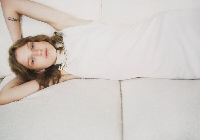 Model lying on a white sofa wearing a sleeveless white dress and a chunky silver chain necklace.