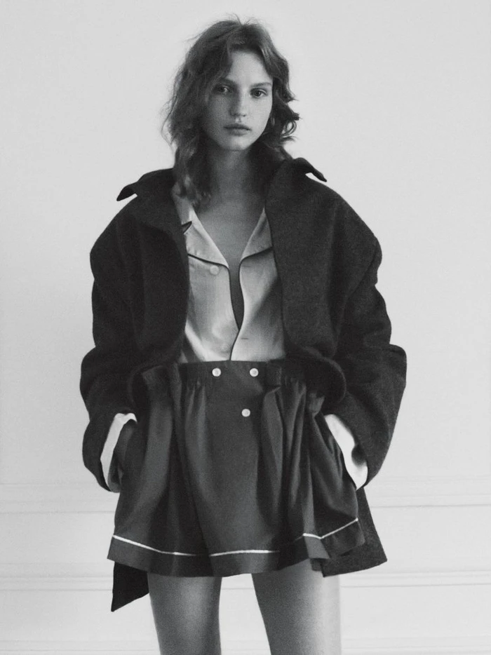 Monochrome fashion editorial portrait of a woman wearing a dark coat and shorts against a white wall.