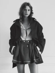 Monochrome fashion editorial portrait of a woman wearing a dark coat and shorts against a white wall.