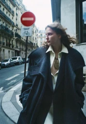 Model Nadine Kirilova wears an oversized dark coat over a cream shirt while walking along a Parisian street near a no-entry road sign.