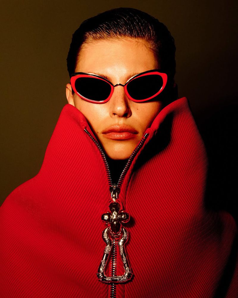 Woman in red sunglasses and matching coat poses indoors with dramatic lighting; beauty and fashion editorial with dark green background.