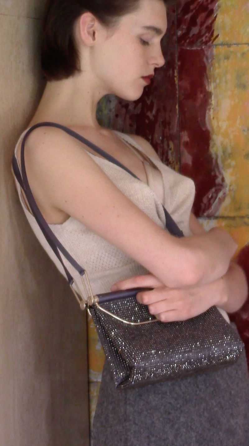 Adult female in gray skirt and knit top poses indoors, holding a sparkly handbag with metallic straps, against a mottled wall.