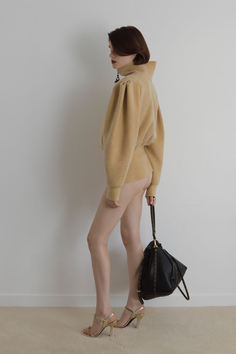 An adult female fashion model poses in a studio. She is wearing a light-brown sweatshirt, high heels, and holding a black purse.