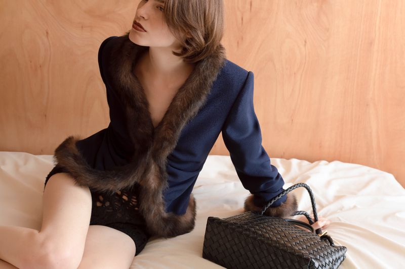 A woman in a fur-trimmed jacket poses on a white bed with a woven leather handbag against a light brown plywood wall background.