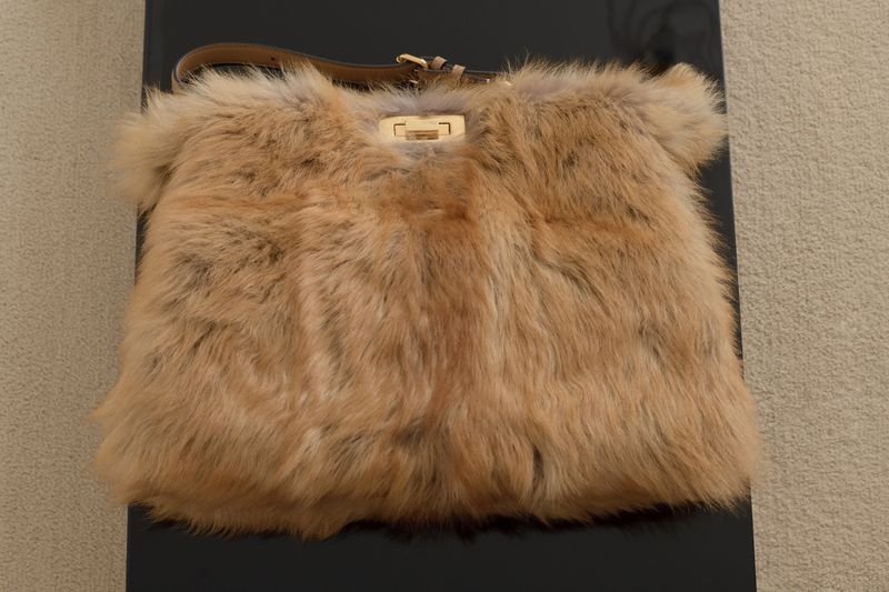Tan fur handbag with brown leather handle and gold hardware on a shiny black surface against a beige background in a studio.