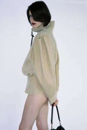 Woman with dark hair poses in a tan sweater and black purse against a white background, side view, fashion shot.