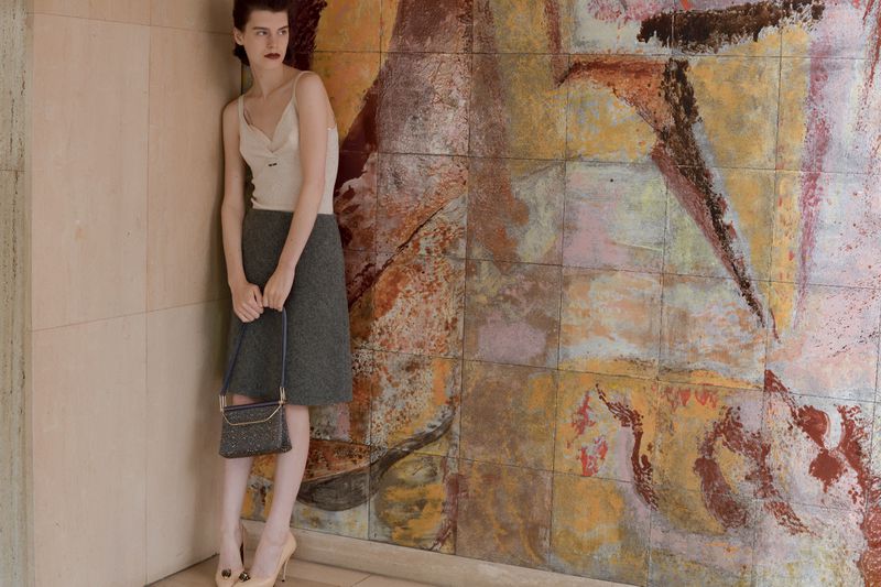 Fashion editorial shot of a slender adult woman with dark hair, wearing a skirt and top, holding a purse against a colorful tiled wall.