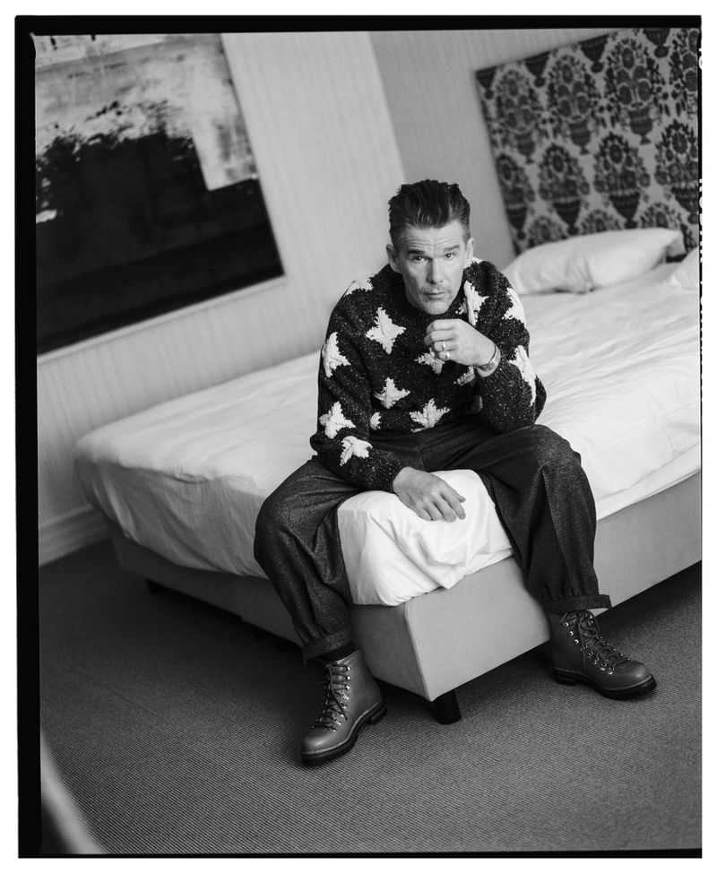 Black and white portrait of a man with styled hair wearing a patterned sweater sitting on a bed in a minimalist room setting.