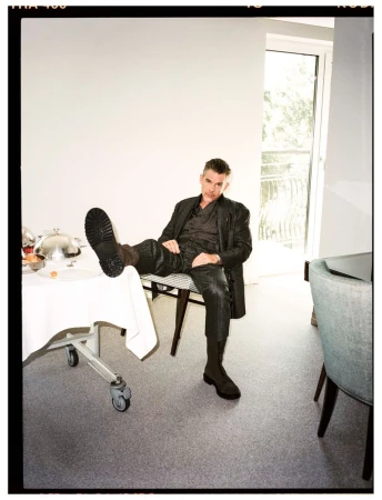 Man in dark suit and boots lounges on a chair, foot resting on a table set for a meal in a light-filled indoor space.