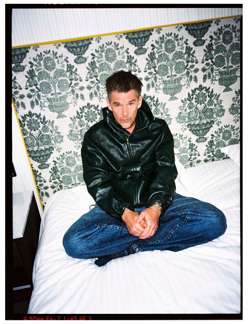 Actor Ethan Hawke sitting cross-legged on a white bed wearing a dark leather jacket and blue jeans in front of a patterned headboard.
